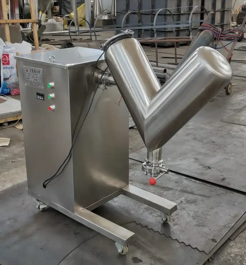The image shows a large stainless steel V-shaped blender or mixer inside what appears to be an industrial manufacturing facility or workshop. The V blender has two cylindrical mixing chambers joined in a V configuration and is mounted on a movable stainless steel frame or stand. Various pipes, hoses and other equipment can be seen in the background of the cluttered workspace. V Blender